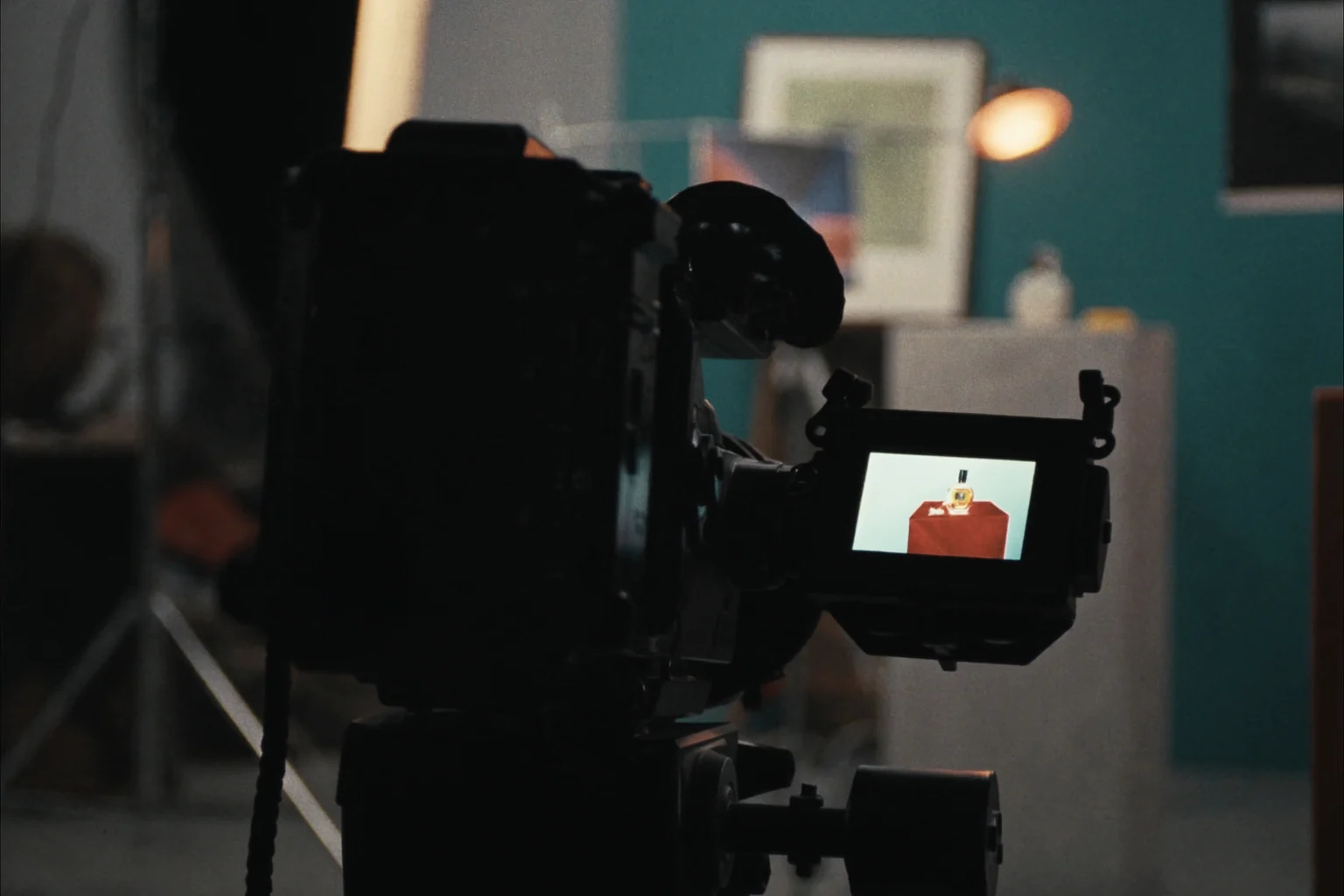 A cinema camera on a tripod in a dim studio, its side screen showing a small product on a red stand.