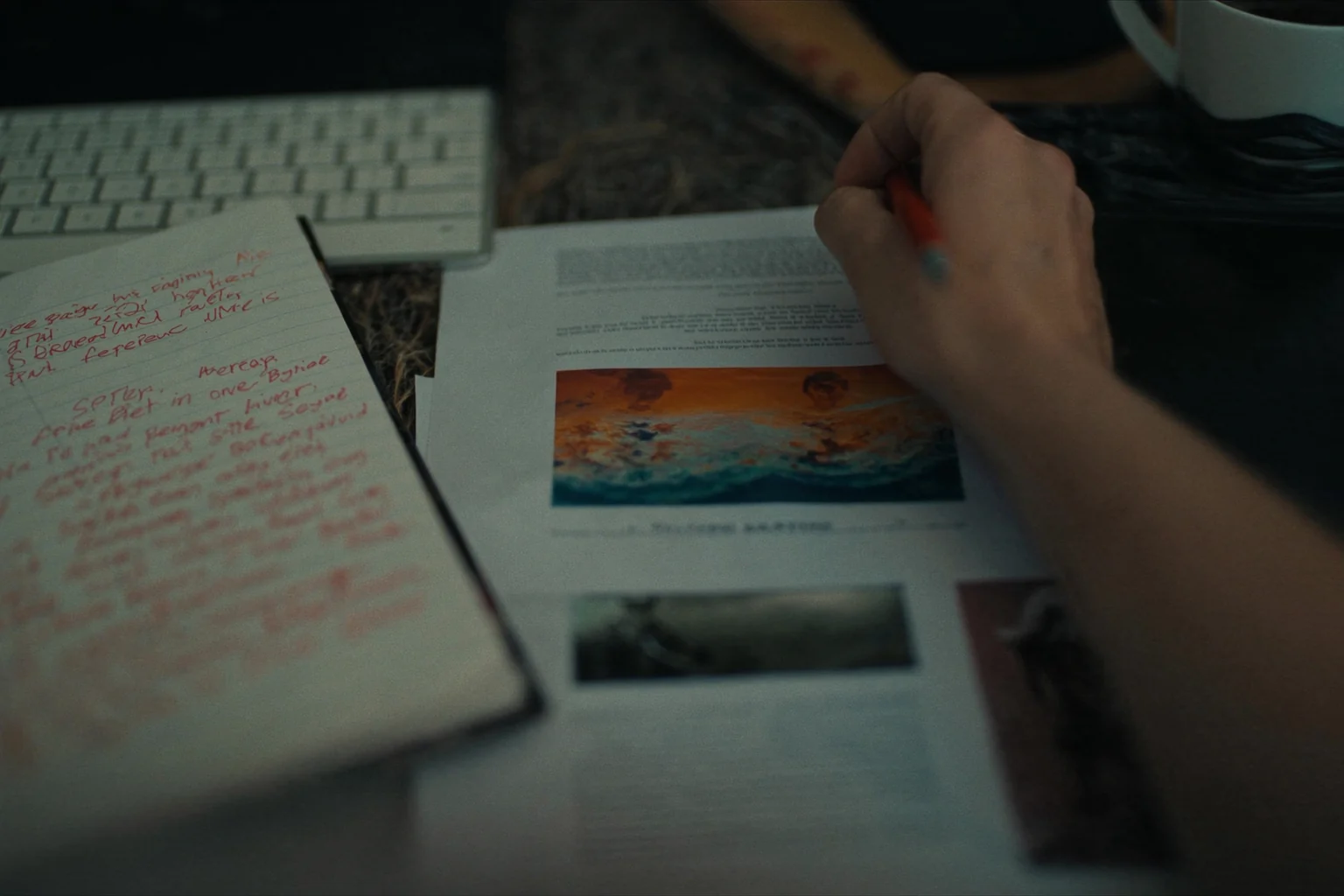 A hand with a red pen marks up printed pages with a picture of a wave, next to a notebook full of red handwriting and a keyboard.