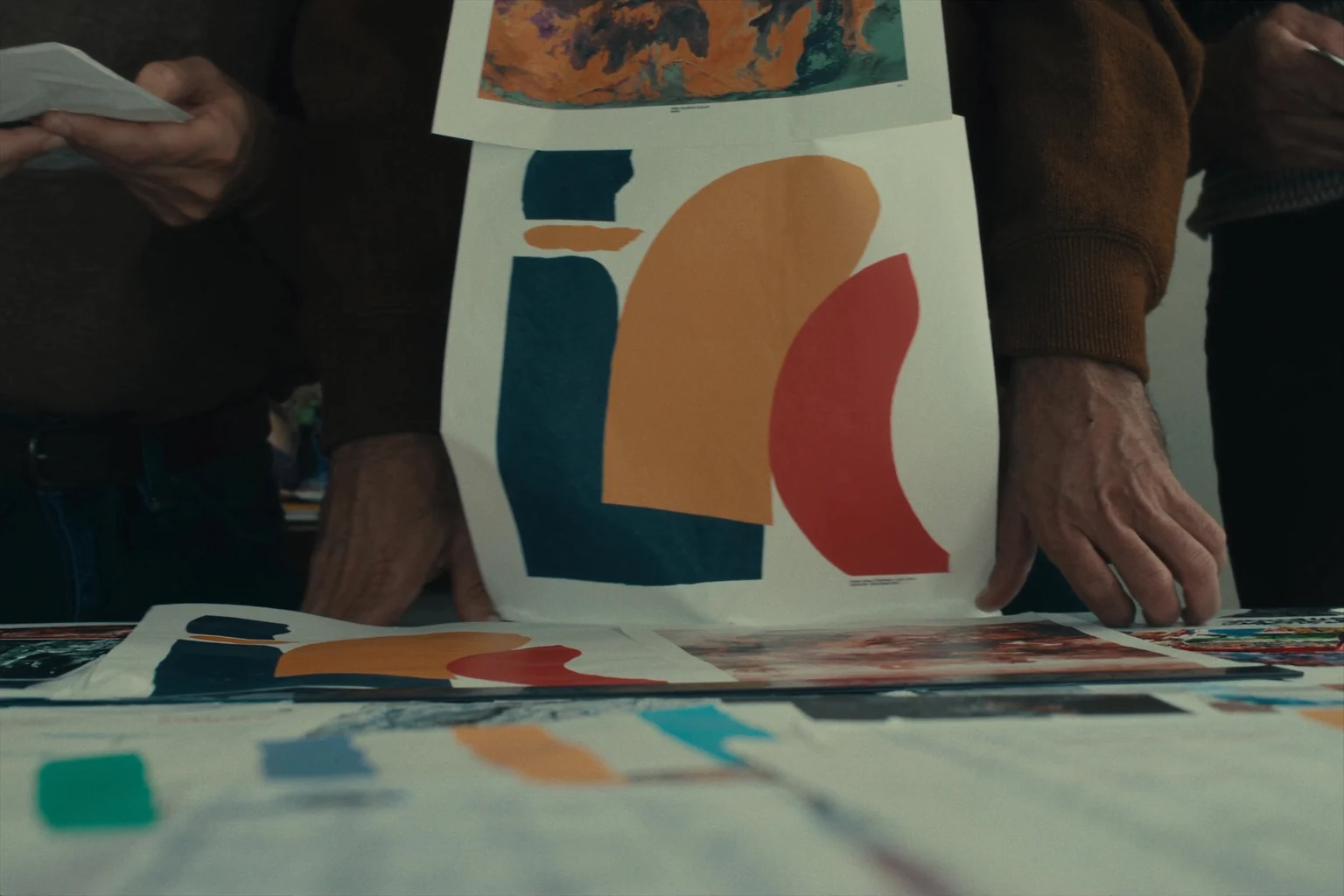 Hands hold up a large printed sheet of teal, orange and red shapes above a table covered with more prints.