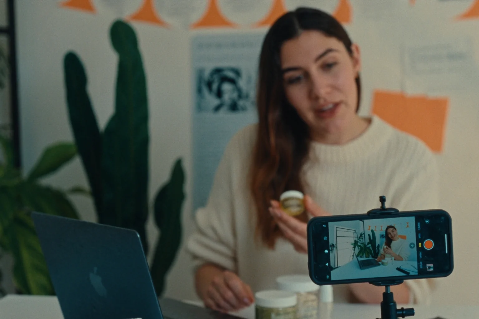 A woman holds up a small jar and talks to a smartphone on a tripod that is recording her, with plants and posters behind her.