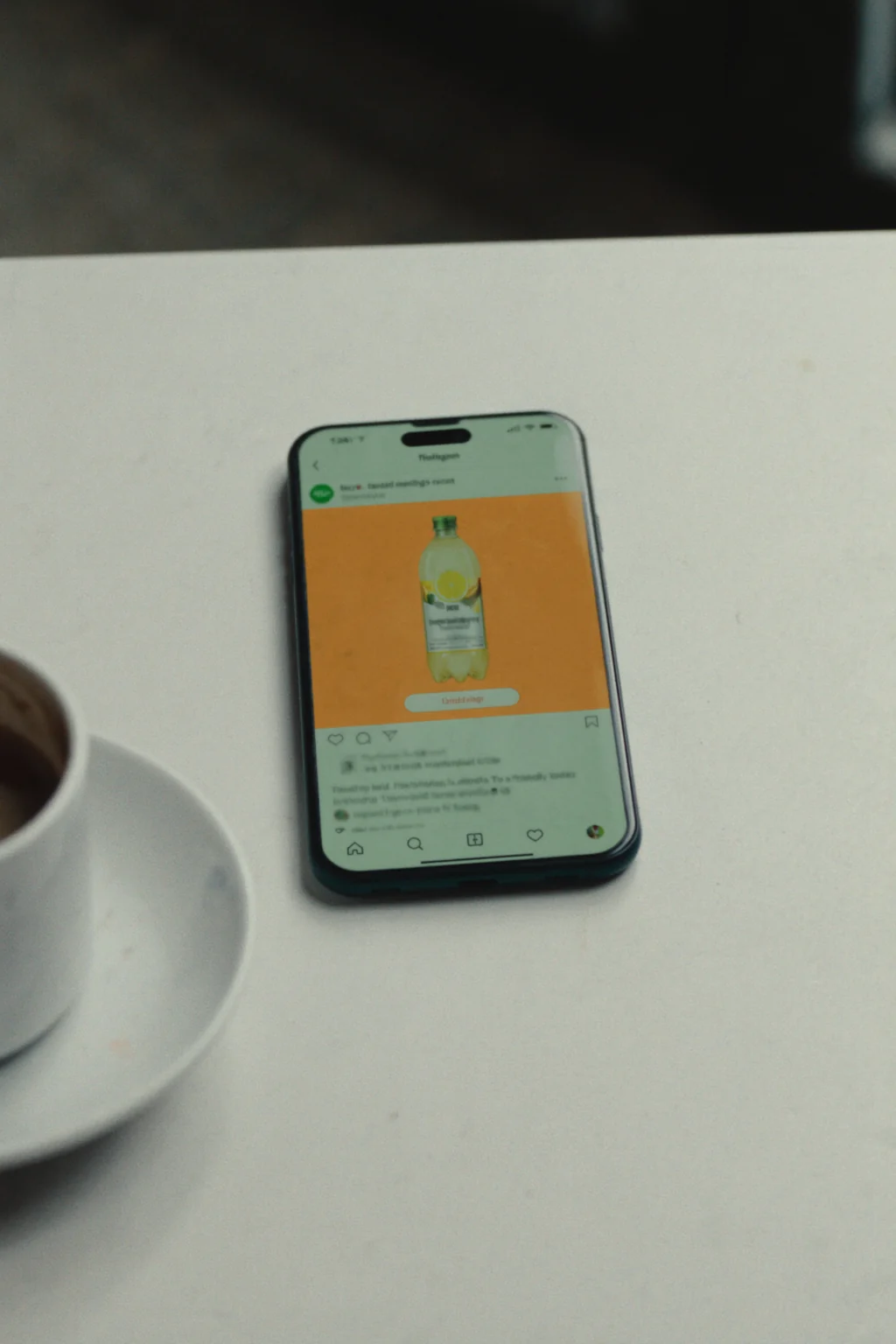 A smartphone on a white table shows a social media post of a bottled drink on an orange background, beside a coffee cup.