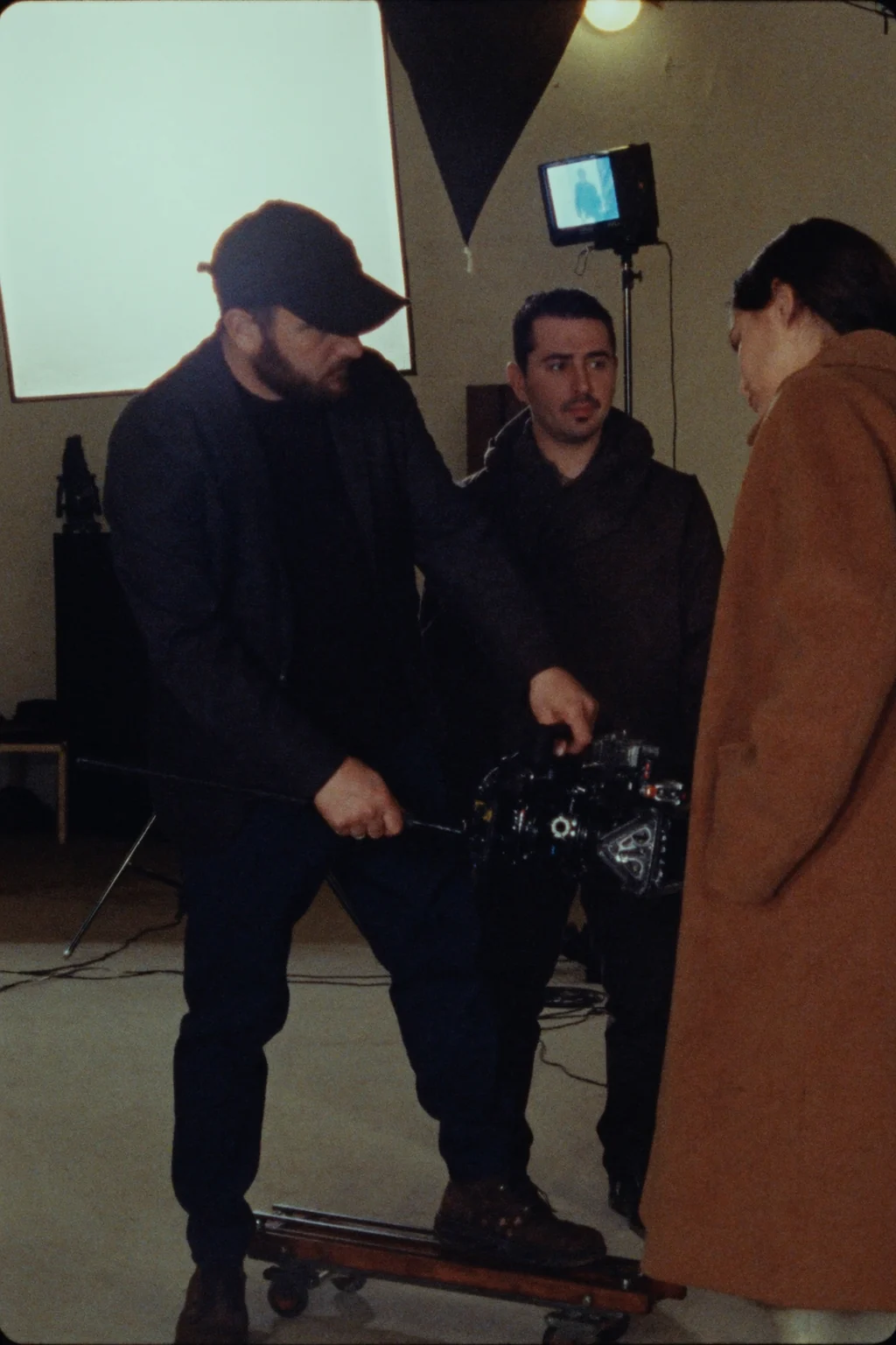 A camera operator in a cap holds a cinema camera in a studio, one foot on a low wheeled dolly, with two other people beside him.
