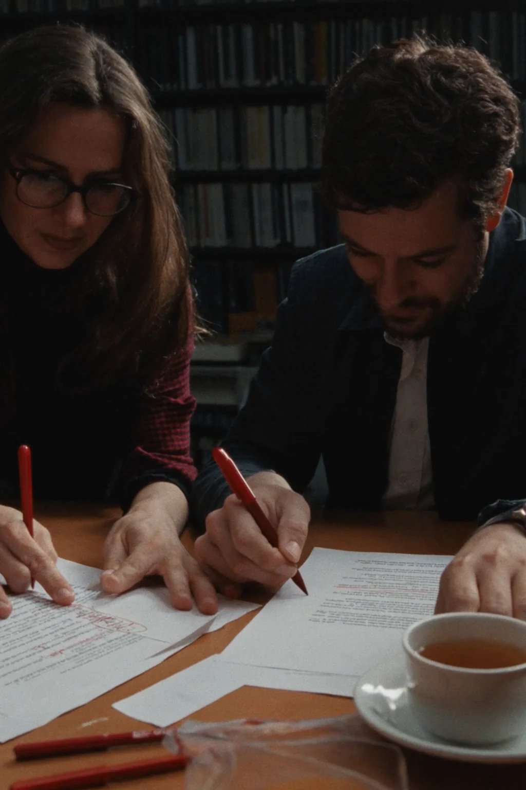 Two people at a wooden table mark up printed pages with red pens, with a cup of tea beside them and bookshelves behind.