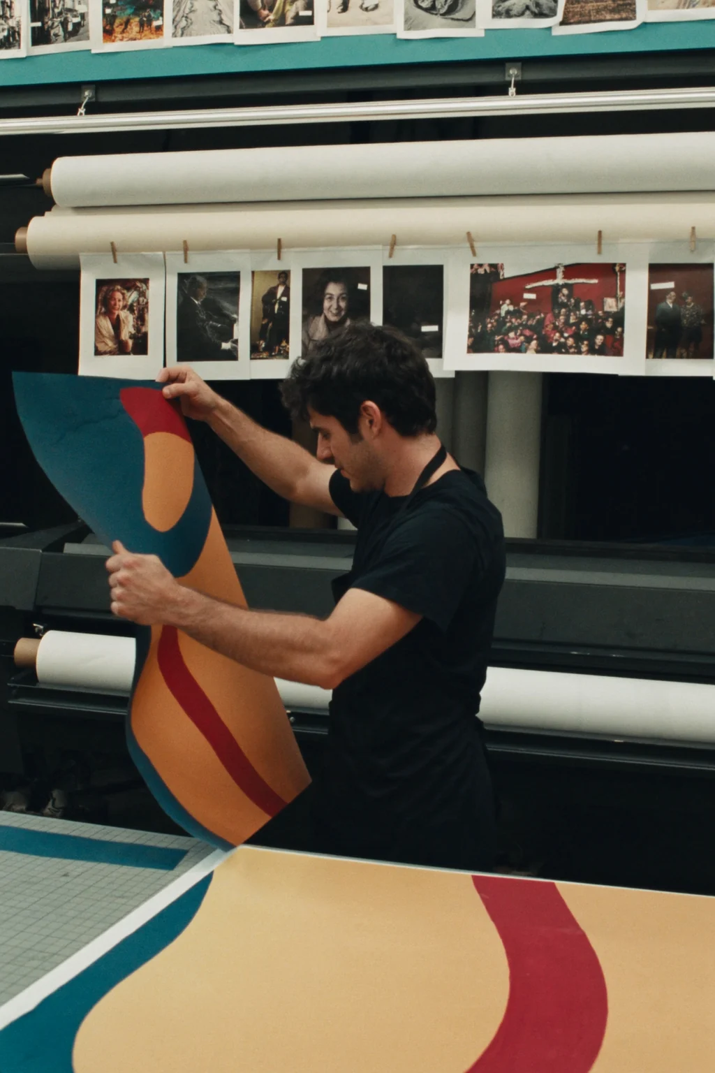 A man in a black T-shirt lifts a large printed sheet of curved teal, orange and red shapes beside a wide-format printer.