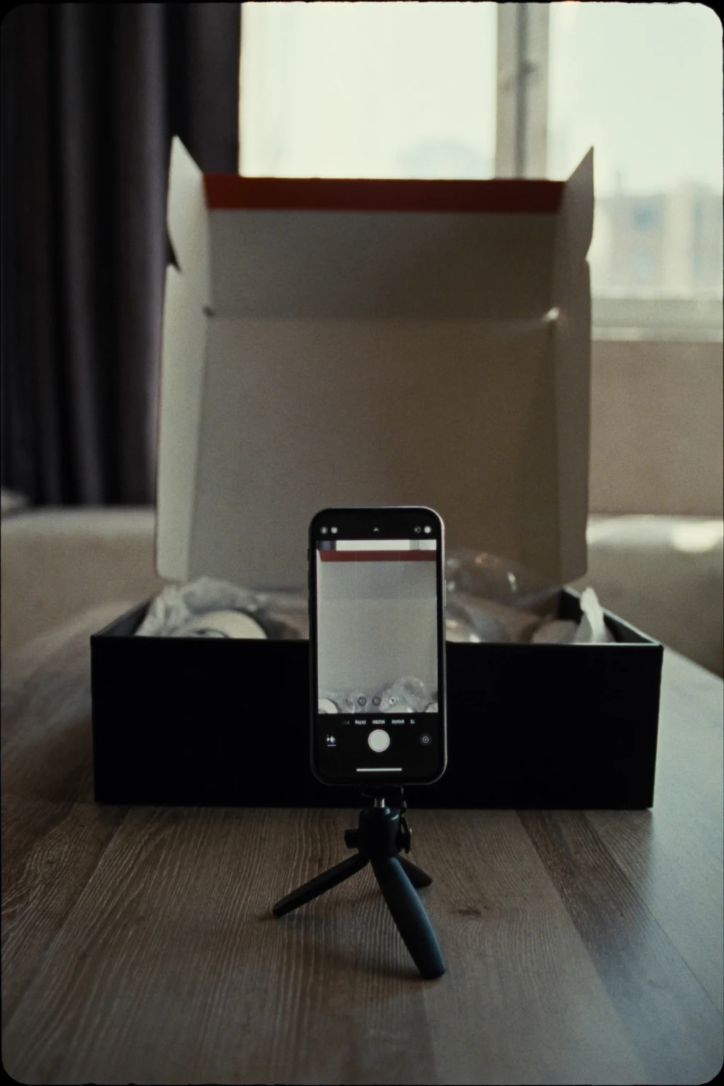 A smartphone on a mini tripod filming an open cardboard box on a wooden table, set up like an unboxing video.