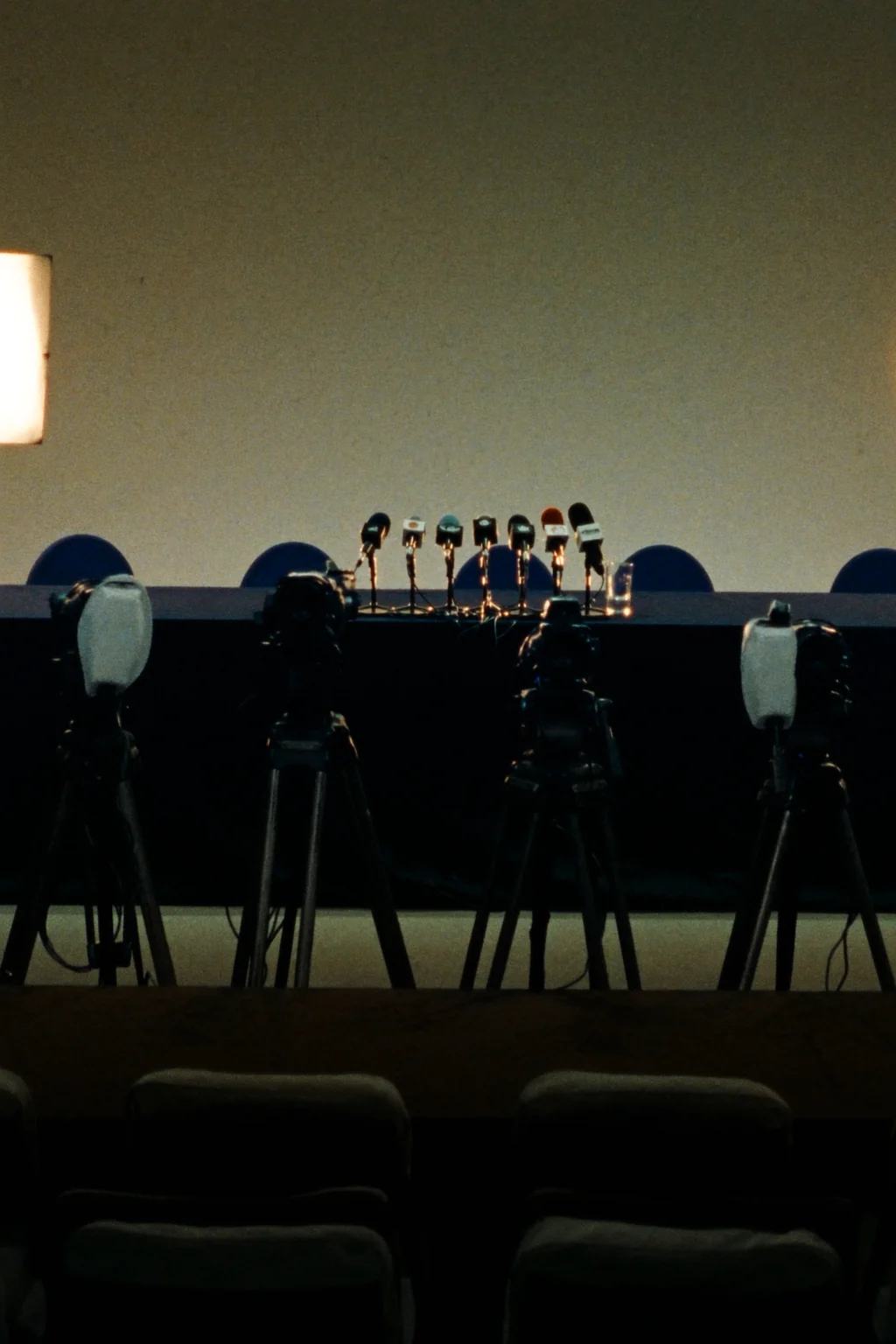 An empty press conference room in low light, with a row of microphones on a long table and camera tripods facing it.