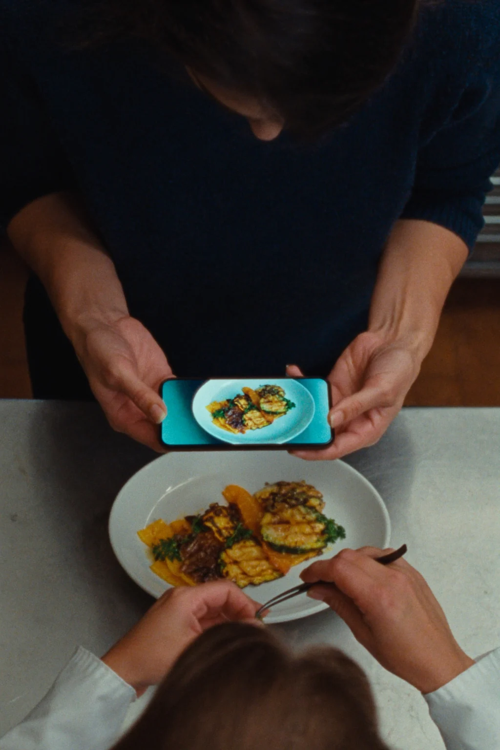 A person photographing a plate of grilled vegetables with a phone from above, while another holds a fork over the dish.