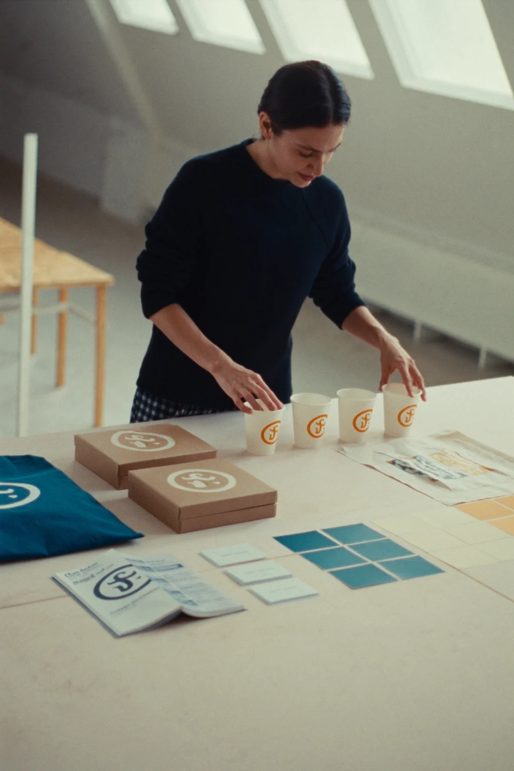 A designer arranging branded paper cups, cardboard boxes, a tote bag and color swatches on a long table in an attic studio.
